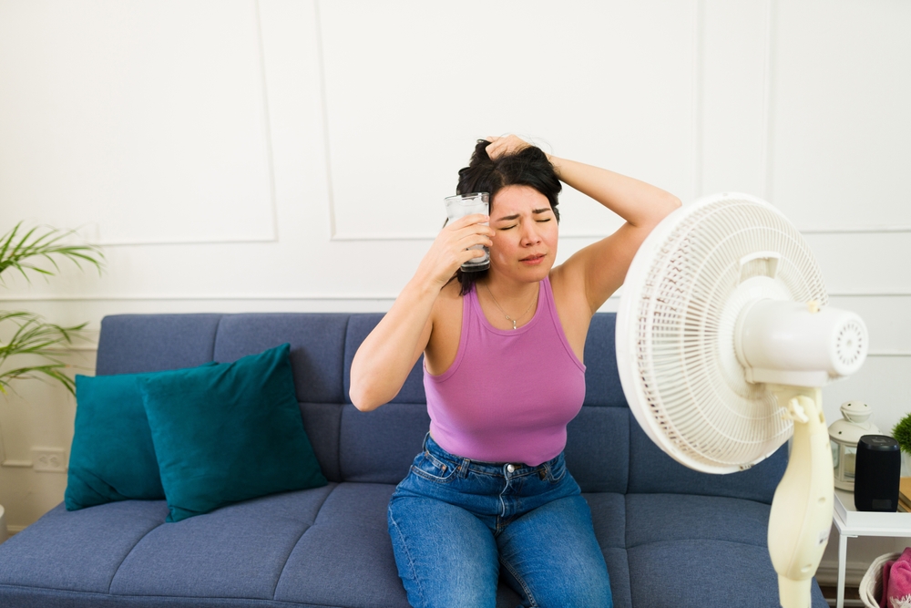 woman holding a glass of ice water to her head as a fan is pointed on her because her AC is running but not cooling